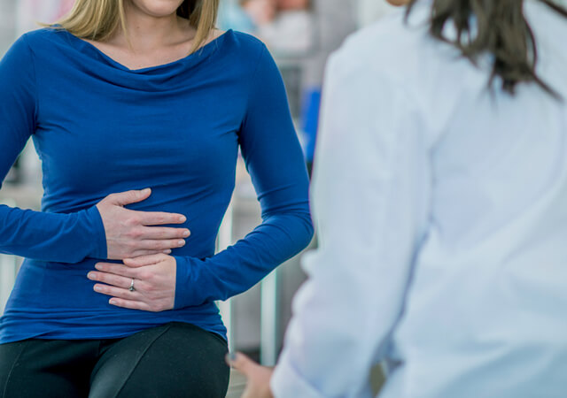 Mulher segura o abdómen enquanto conversa com um médico, representando consulta médica para dor abdominal.