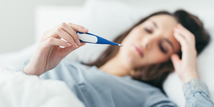 Mulher deitada na cama, com uma expressão de mal-estar, a segurar um termómetro e com a mão na cabeça.