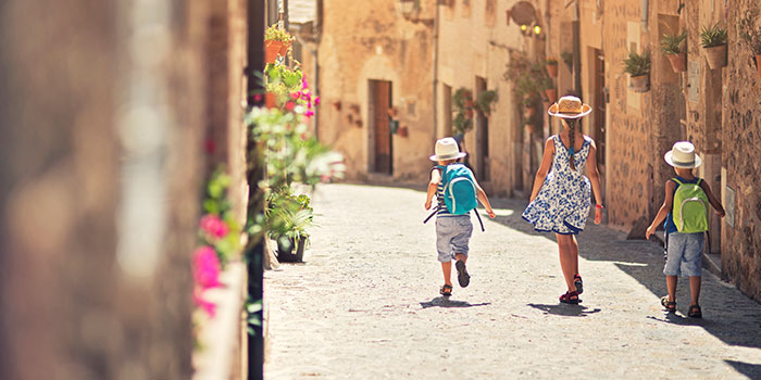 Três crianças caminham numa rua de paralelepípedos numa vila histórica, com chapéus e mochilas, rodeadas por vasos de flores e iluminadas pelo sol.