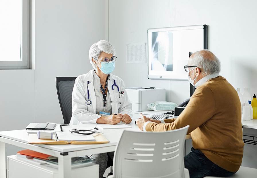 Médica no consultório, à conversa com paciente idoso durante uma consulta médica, enfatizando a importância da prevenção e diagnóstico precoce do cancro.