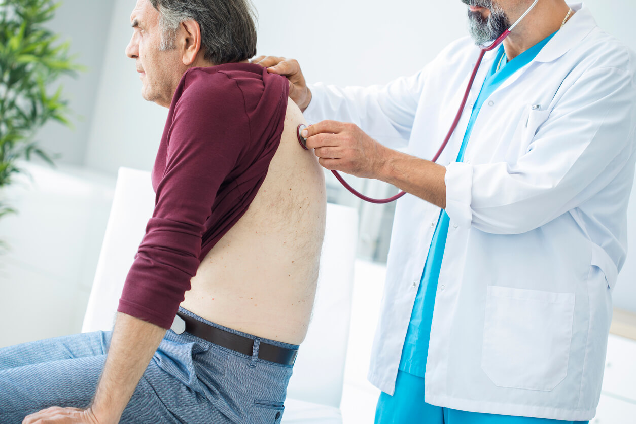 Profissional de saúde a auscultar as costas de um paciente com um estetoscópio em um consultório médico, para analisar uma possível pneumonia.