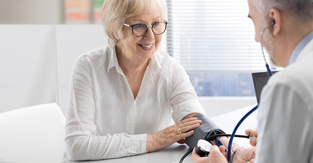 Paciente idosa a sorrir durante uma consulta médica enquanto o médico mede a sua pressão arterial, ao fazer o diagnóstico da cardiomiopatia.