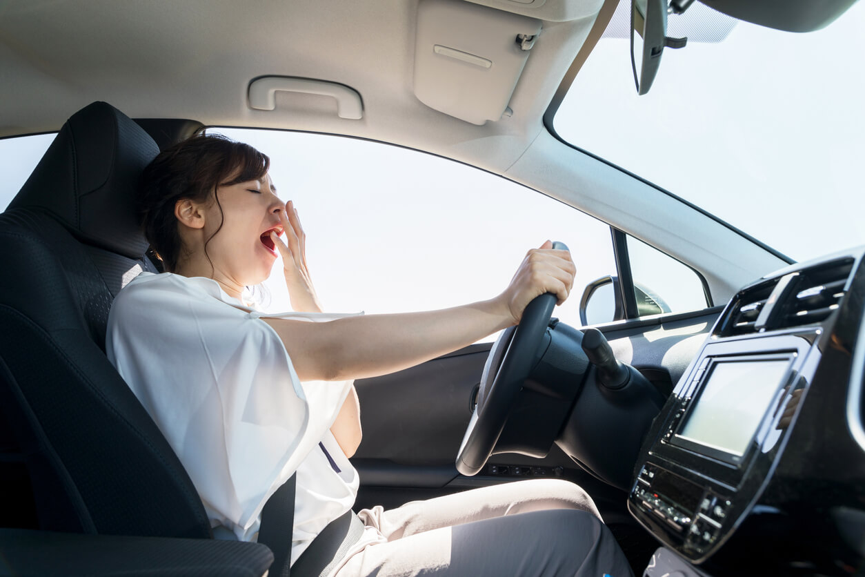 Mulher ao volante do carro com a mão à frente da boca, sinal de quem está a bocejar. Isto destaca a importância de uma boa noite de sono para a nossa saúde